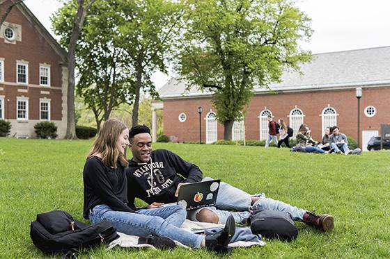 Two Chatham University students sit on a green grass 学术 quad, working toge的r on a laptop. 