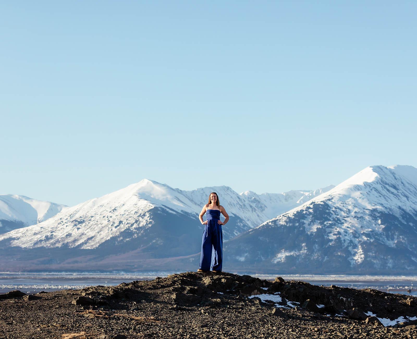Portrait of Chelsea Kovalcsik standing outside with her arms on her hips, in front of a beautiful Alaskan mountain on a sunny day with a bright blue sky